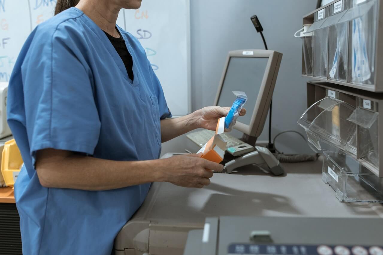Person in scrubs handles medical equipment near a computer station.